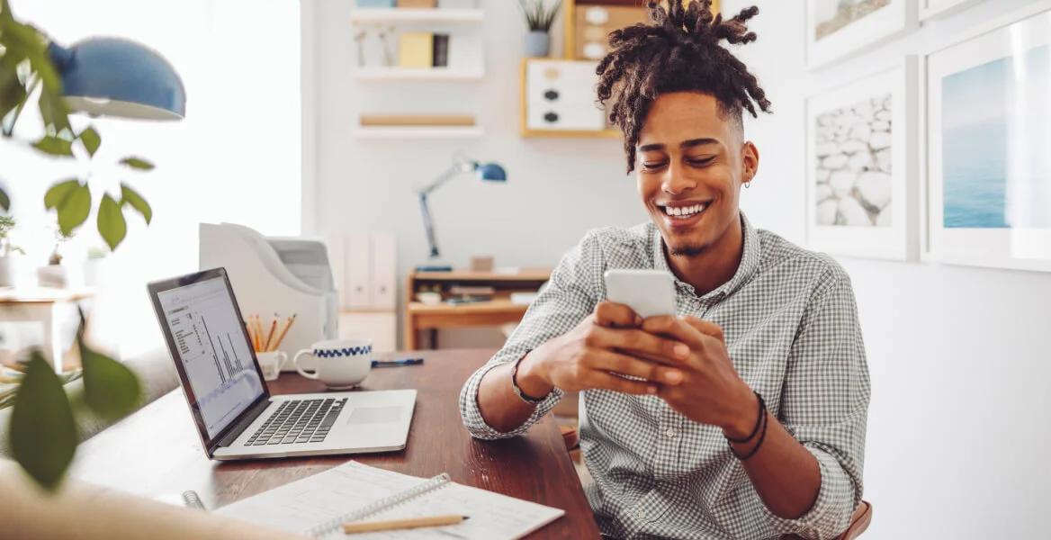 Jovem sentado e sorrindo enquanto mexe no celular.